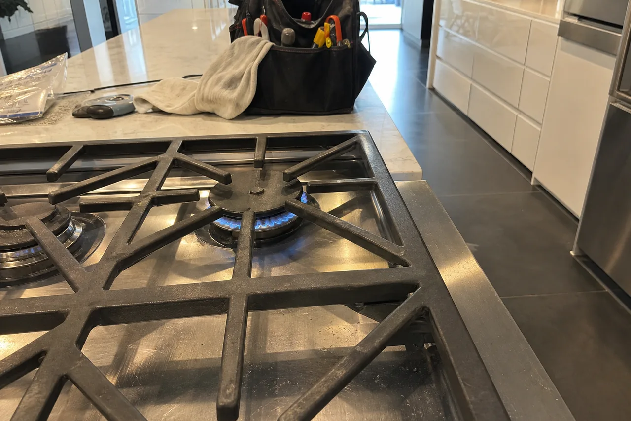 A low blue flame burning under heavy cast-iron grates on a stainless gas rangetop, with a tool bag and a gray rag resting on the marble island behind it