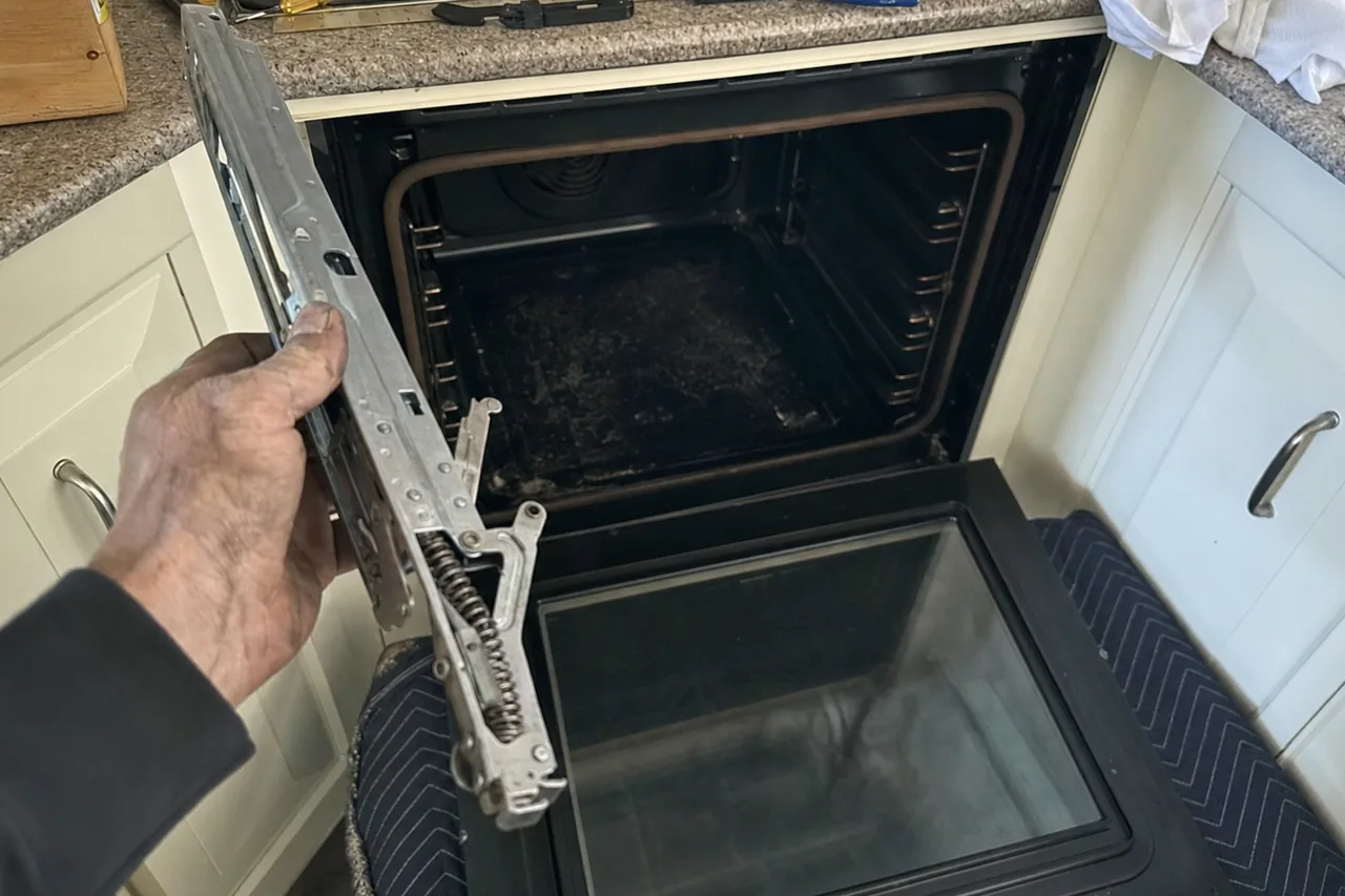 A hand holding an oven door hinge with its coil spring exposed, the door glass resting on a chair